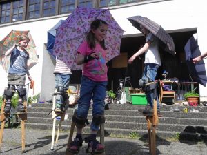 Foto: Kinder tanzen auf Stelzen mit aufgespannten Regenschirmen
